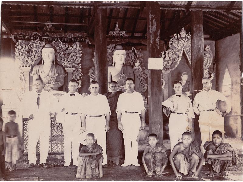 Six International Drillers wearing white inside a temple with five young boys and a monk. There are three pillars running from the floor to the ceiling and three statues in the background.
