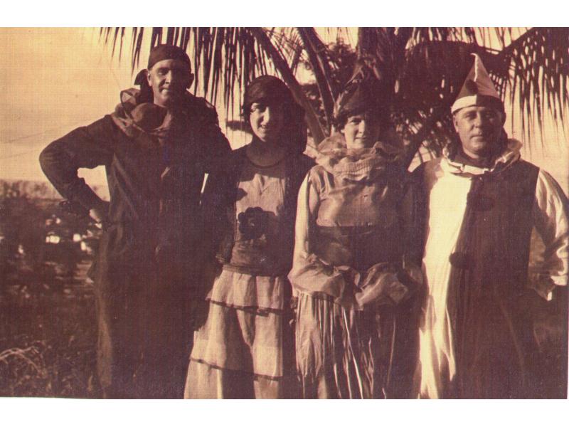 Two adult couples in costume, on their way to a masquerade party. The couple on the left appear to be pirates and the couple on the right, clowns. They are standing in front of a palm tree.