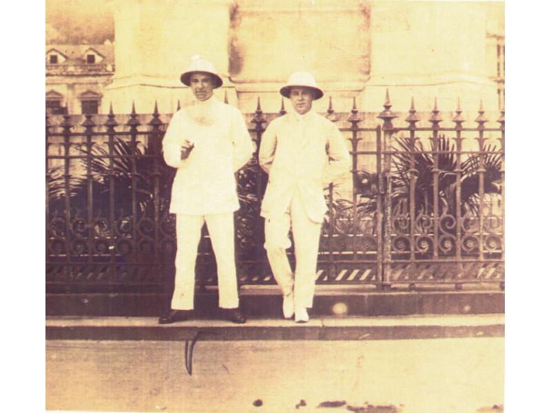 Two drillers dressed in white suits and pith helmets standing against an iron fence. There is horse manure in the street. 