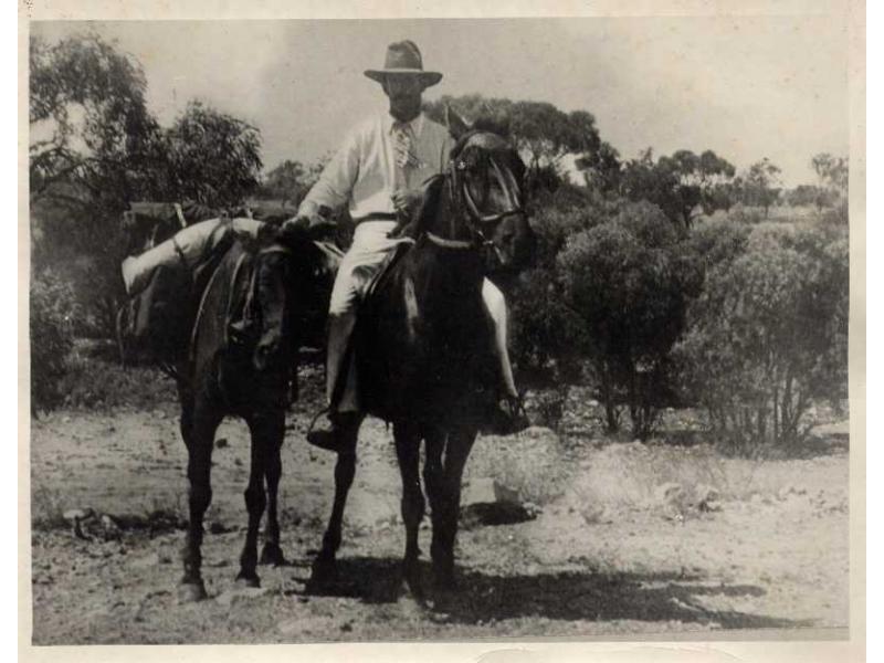 William Gillespie sitting on a horse and holding another horse that is loaded with baggage. He is wearing white clothes and a hat. There are trees behind him.