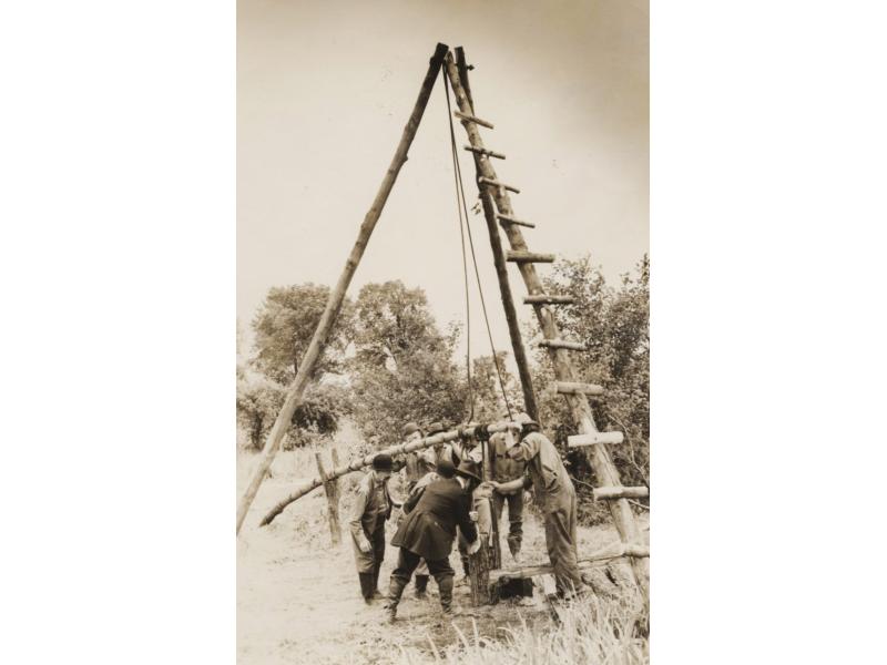 Un groupe d’hommes est réuni autour de la base d’un mât basculant, installé sous un derrick à trois mâts.