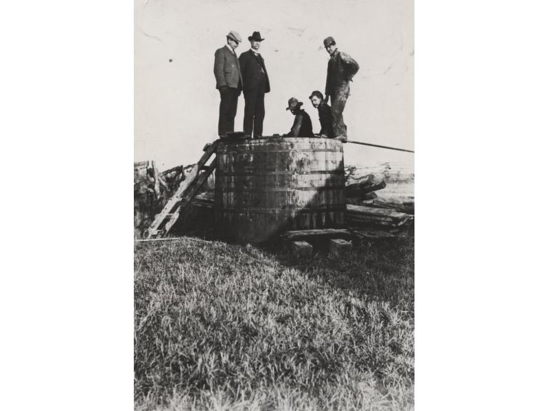 A group of five men standing on an above-ground wooden storage tank. There are metal rings around the barrel holding it together. There is a ladder propped against the tank so the men could climb on top of it.