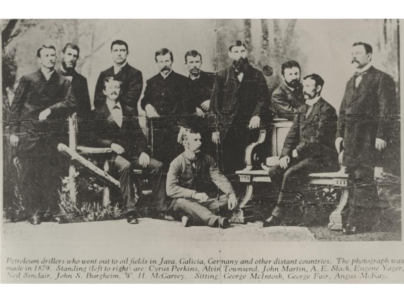 A group of men in front of a painted background. They are wearing dark jackets. Seven are standing, three are sitting in chairs, and one is sitting on the floor.