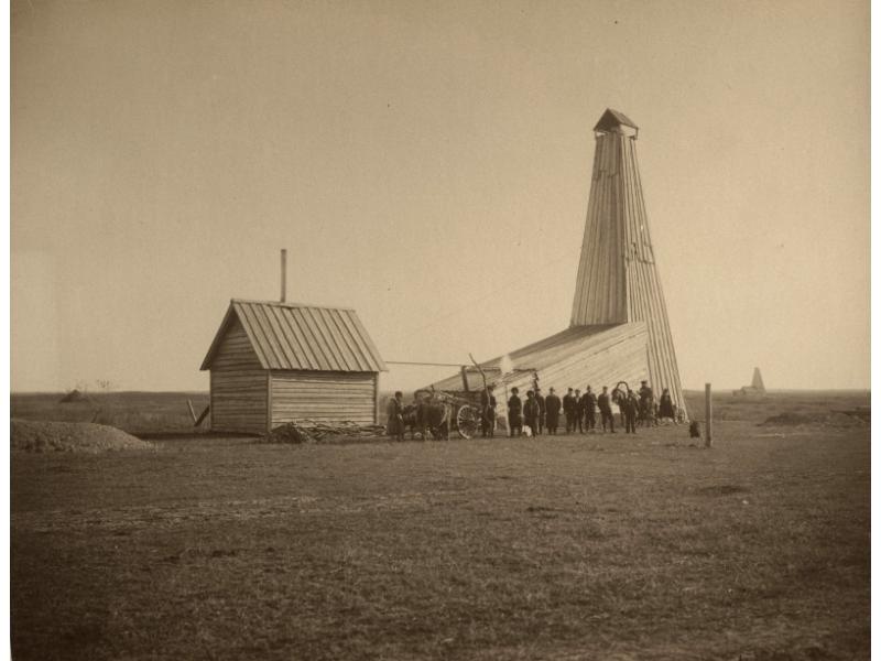 A group of people standing in front of an oil rig that has been covered in wooden planks. They are in a flat field and there is another rig in the background.