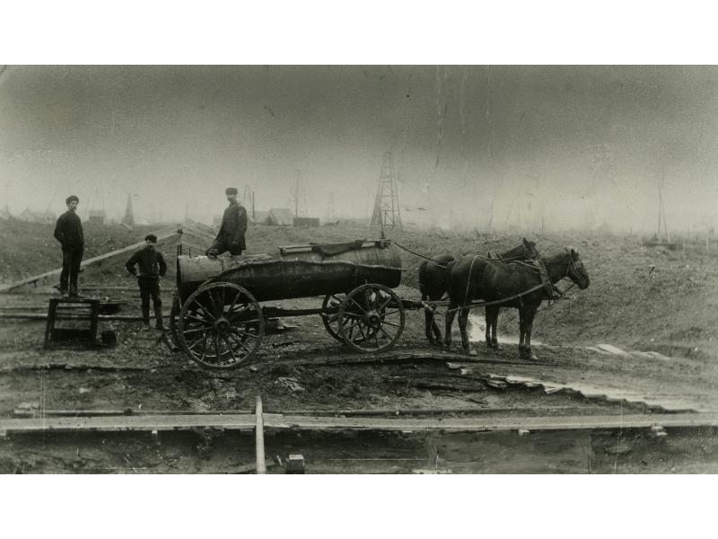 Two horses are harnessed to a cylindrical tank wagon with four wheels. Three men stand behind the wagon and there are oil rigs in the background.