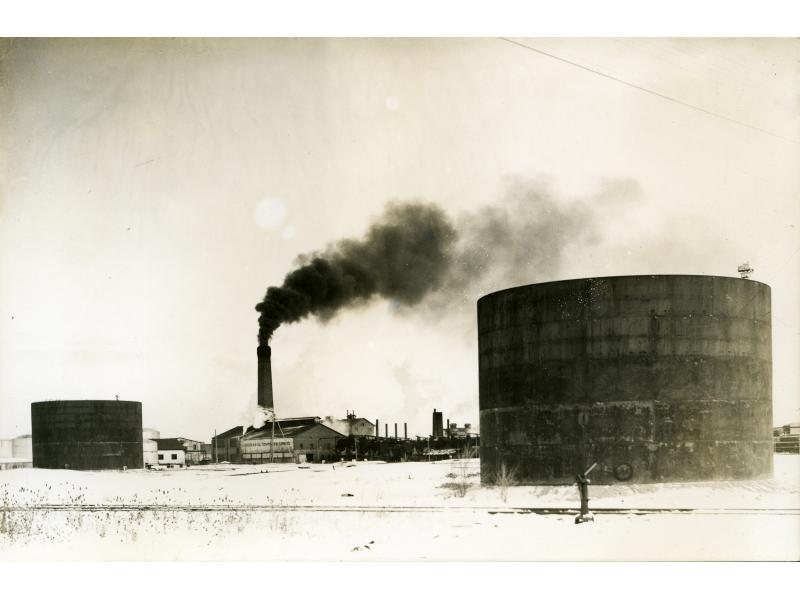 Une photo de deux grands réservoirs de stockage cylindriques de surface, il y a des bâtiments et une cheminée produit de la fumée noire derrière eux. Une voie ferrée traverse l’avant-plan.