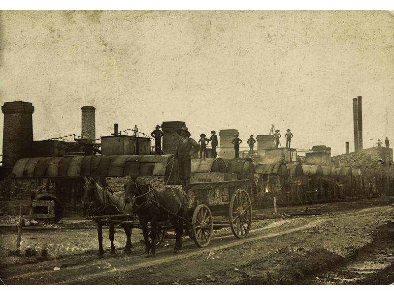 A man stands on a cart pulled by two horses on a dirt road. Behind him, a group of men stand on small buildings with curved roofs that have chimneys coming out of the tops.