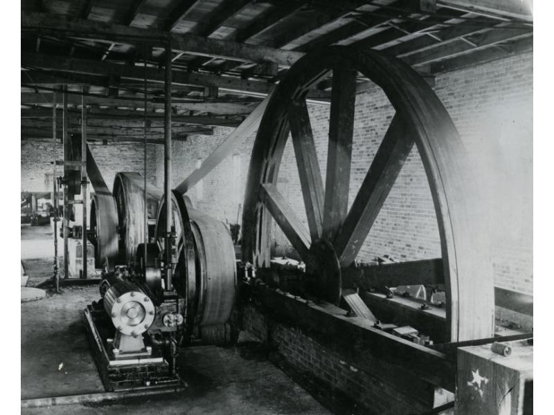 A pumping rig with a large wheel and three smaller wheels inside a building. There are belts that loop around the wheels. There is a motor in the foreground.