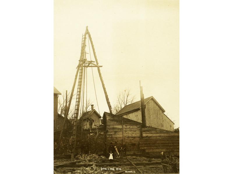 Un derrick à trois mâts installé par-dessus une installation de forage à Petrolia. Un homme se tient sur l’installation de forage et des hommes sont autour de la base. Une fillette et un chien sont aussi au bas.