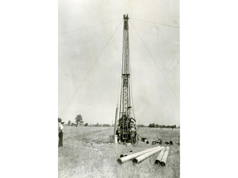 A photo of a small drilling rig in a field. There are four wires extending from the top of the rig down to the ground. Three men are looking at the rig and there are pieces of casing laying on the ground in front of it.