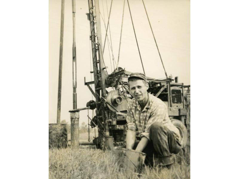 A photo of a man with his hands in a bucket, in front of a pulling rig that has three visible posts and wires running from it to the vehicle carrying it.