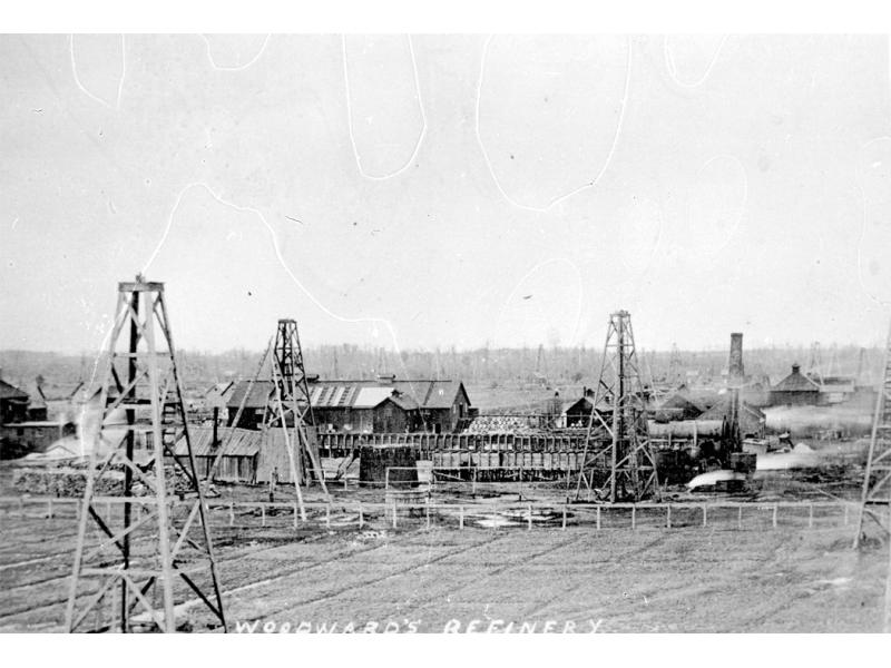 A field with three oil derricks in it. There are buildings in behind with pipes running between them. There are more derricks in the background. Along the bottom of the photo, "Woodward's Refinery" is written in white.