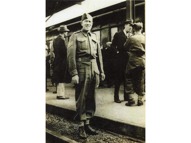 A man wearing a military uniform, standing beside railway tracks in front of a train. There are people standing on the platform behind him.