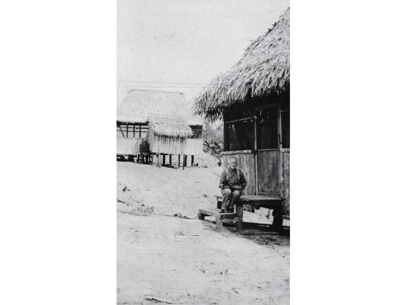 A man sitting on the steps leading up to a thatched building on stilts. There is another building in the background.