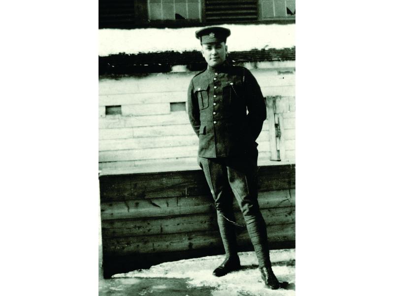 Bloss Sutherland standing outside a building with wooden siding. There is snow on the ground and he is wearing his military uniform and hat.