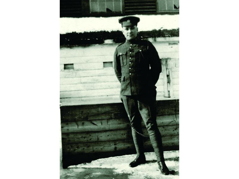 Bloss Sutherland se tient debout à l’extérieur d’un bâtiment au revêtement de bois. Il y a de la neige au sol et il porte son uniforme militaire et un chapeau.