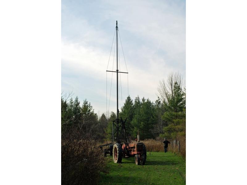 A photo of a portable pulling rig set up in a grassy field, pulled by a red tractor. It has a tall beam with a shorter beam running horizontally, like a cross. A man leans on a fence behind it.