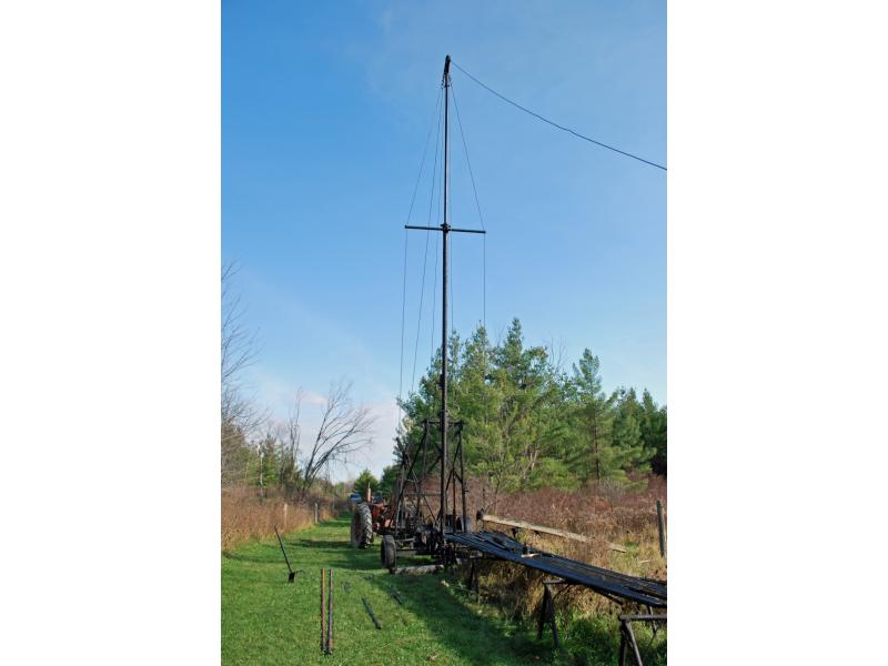 A photo of a modern portable pulling rig, with cables extending down to the pump. It has a tall beam with a shorter one running horizontally, like a cross. There are metal poles lying on wooden saw horses in front of the rig.