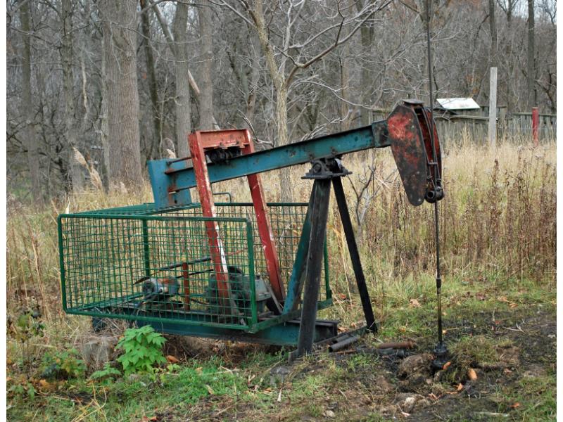 A nodding-head pumping rig powered by a diesel engine, painted green and red. There is a metal basket over the engine. There is a metal line running from the head into the ground.