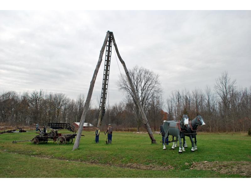 A three-pole derrick with tin figures around it, showing a team of horses, man at the pump head, and a boy and dog sitting on a portable pump kit.