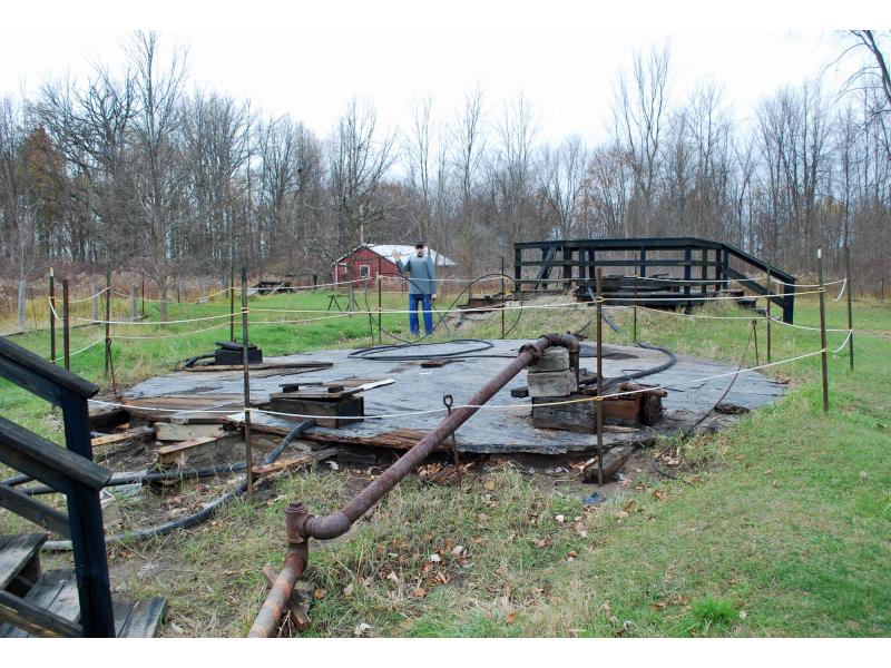 Un réservoir de stockage en bois souterrain, avec un tuyau allant jusqu’à la route. Il y a des poteaux et de la corde autour du réservoir, et une personne se tient derrière, saluant le la main.