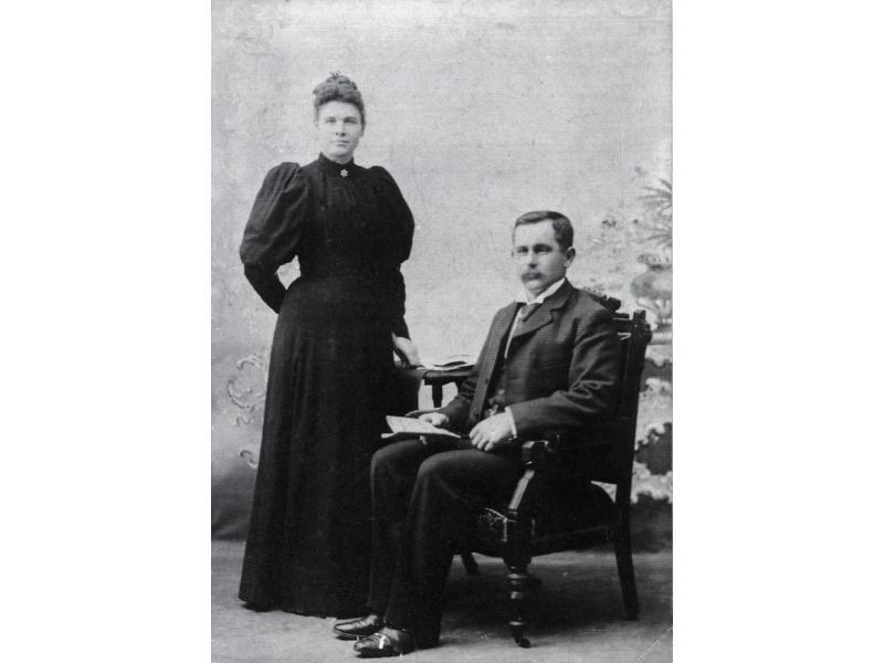 A portrait photo of Elizabeth Douglas and James Boyd in front of a painted background. She is wearing a full-length dark dress with puffy sleeves and a brooch at the collar. He is sitting in an armchair holding a piece of paper and he is wearing a dark suit.