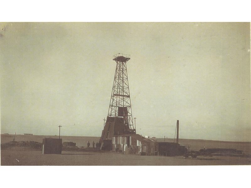 A photo of three men standing beside an oil rig in a flat field. The bottom portion and machinery are covered with pieces of metal.