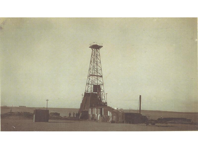 Une photo de trois hommes debout à côté d'une installation de forage pétrolier dans un champ plat. La partie inférieure et la machinerie sont recouvertes de tôle.