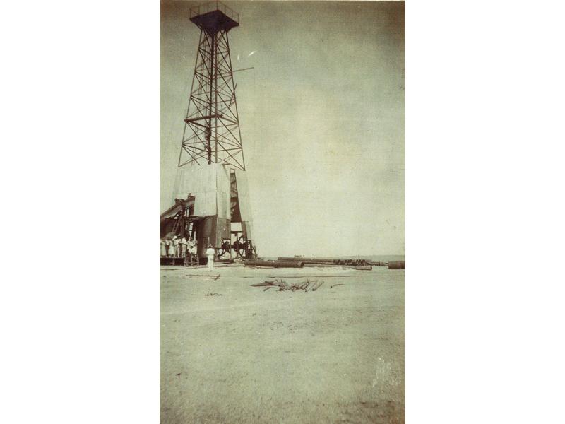 A group of people stand at the base of an oil rig with a platform at the top. The bottom part of the rig is covered with sheets of metal. There is a pile of casing off to the right side.