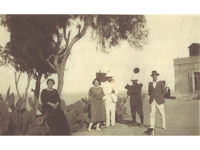 Two women in long dresses standing beside three men in suits and hats. There is a tree behind them and the corner of a stone building is visible.