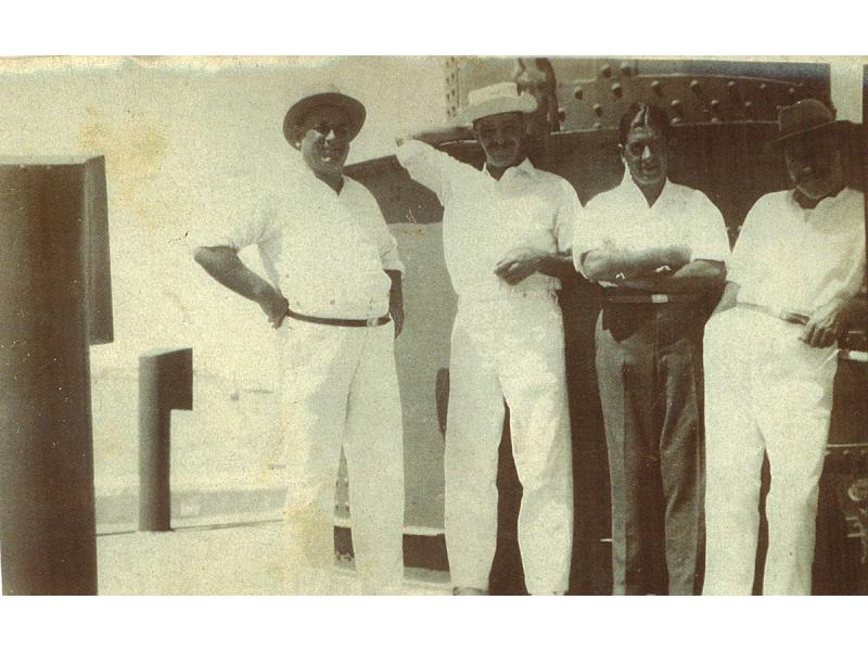  Une photo de quatre hommes vêtus de blanc sur le pont d'un navire. Ils sont debout devant une colonne de métal munie de rivets.