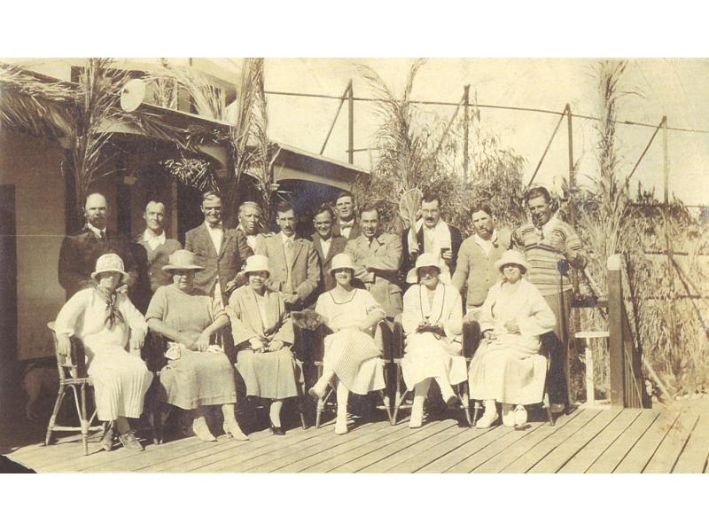 A photo of two rows of people on a wooden deck. The men are standing in the back row, wearing suits. The women are sitting in chairs in the front, wearing dresses and hats