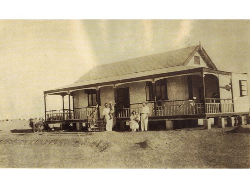 Two men, a woman, and two children standing in front of a small house with a porch. They are all wearing white. There is sand in the foreground and background.