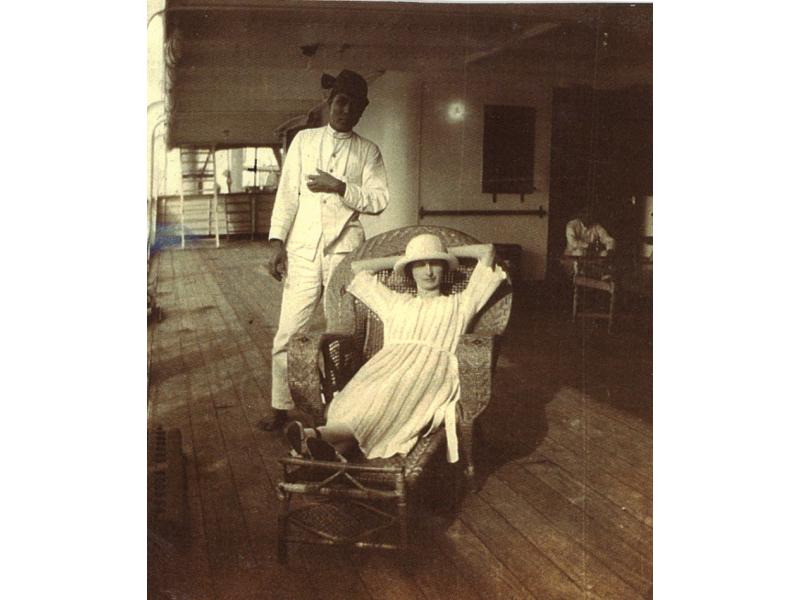 A woman lying back in a wicker reclining chair, wearing a white dress and hat. A man in a white suit stands behind her. They are on a ship deck with a wooden floor.