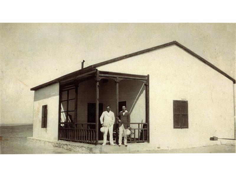 Ross Dyke and another driller standing in front of a white house with a dark railing around the porch, holding white pith helmets.