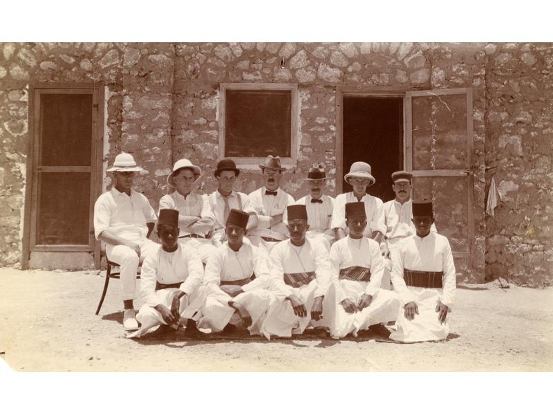 Photographie de six foreurs pétroliers en Égypte, avec cinq membres du personnel domestique. Ils sont assis devant un bâtiment. Les foreurs internationaux portent des costumes blancs et les domestiques portent des robes blanches avec des ceintures en tissu et des chapeaux noirs.