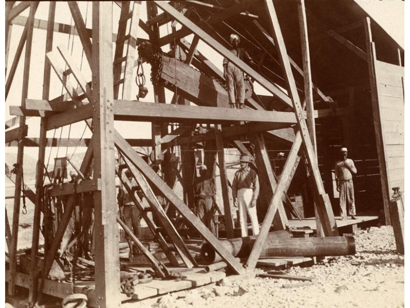 William Gillespie and his workers underneath a wooded rig on Jubal Island. Gillespie is wearing white pants and a pith helmet.