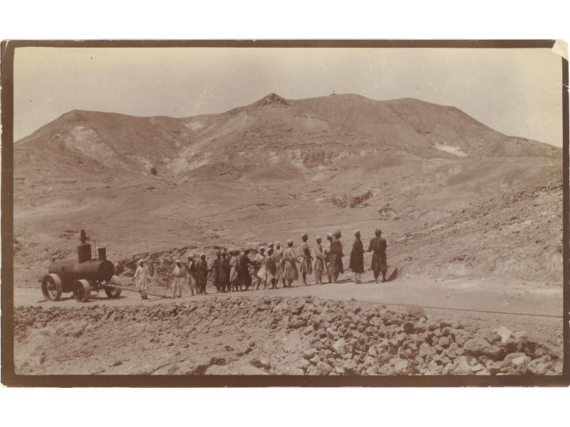 Un groupe d’hommes tire une chaudière avec une corde sur une légère pente dans le désert. Il y a des pierres sur la route et des collines en arrière-plan.