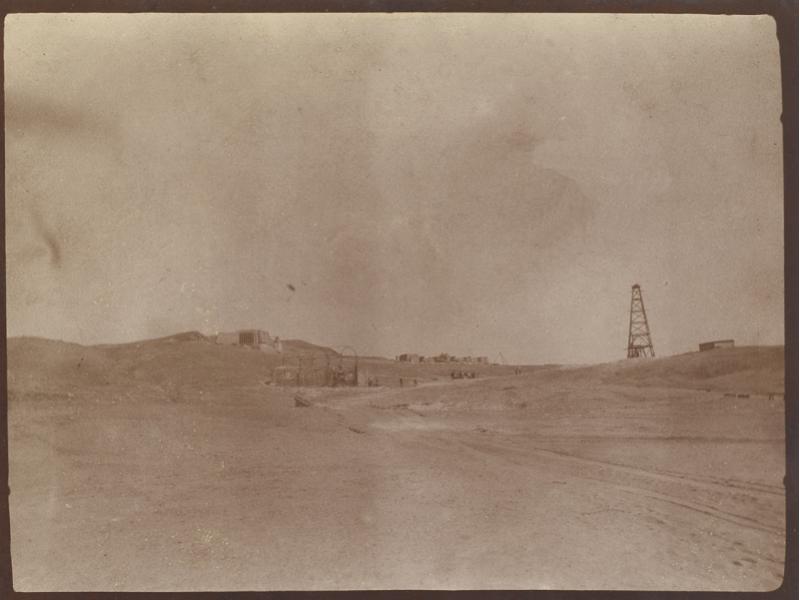 A photo of a barren field with an oil rig on the right. There are two structures on the right and a cluster of buildings in the background.