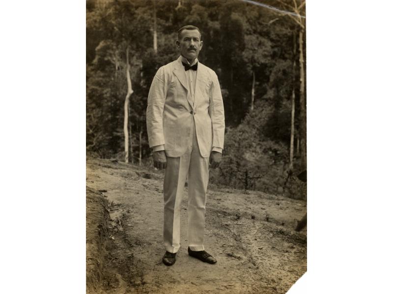 A photo of William O. Gillespie in a white suit with a dark bow tie and shoes. He is standing on a path in front of trees.