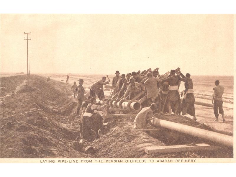 Image en noir et blanc d'une vingtaine d'hommes utilisant de grosses clés comme leviers pour relier ensemble deux gros morceaux de pipeline dans un désert.