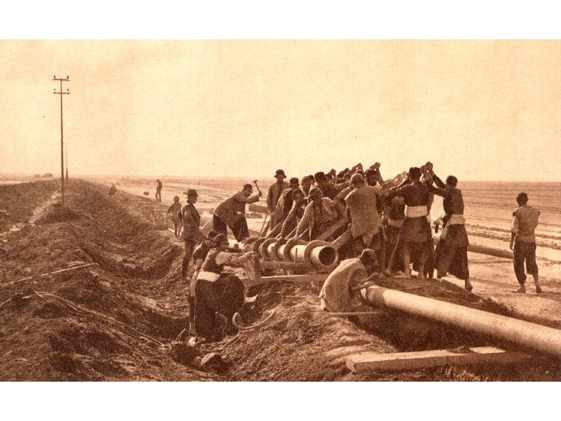 Un groupe d’hommes déplaçant un morceau de conduite dans une tranchée, avec des morceaux de bois et des marteaux. Il y a des poteaux électriques au loin à la gauche.