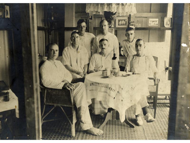 Une photo de sept foreurs internationaux à l'intérieur, autour d'une table avec une nappe blanche. Ils sont vêtus de blanc et quatre sont assis sur des chaises en osier. Une tasse, une pipe et une bouteille sont posées sur la table.