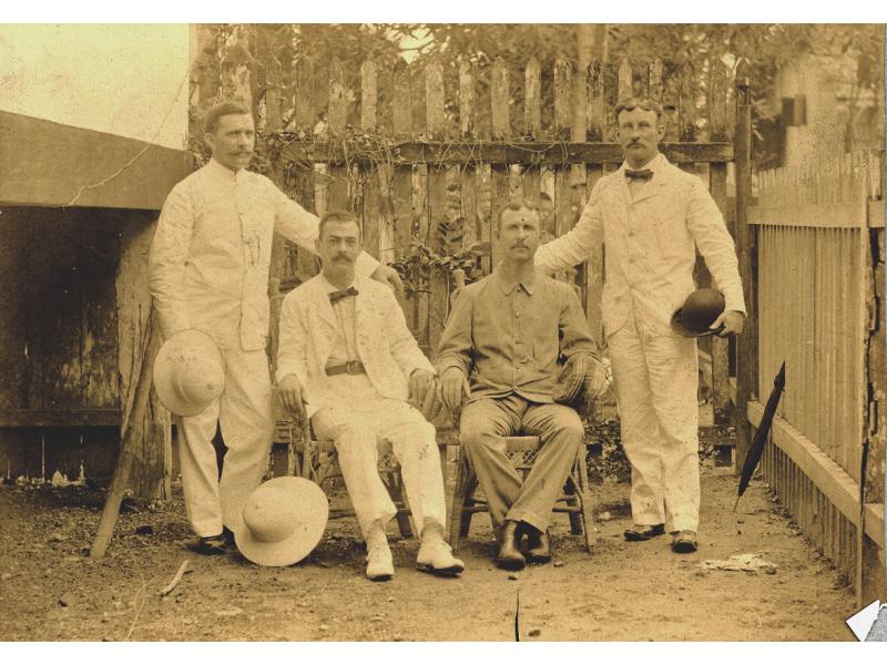 Two men sitting in chairs with a man standing on either side. They are wearing white and light coloured clothing. Each man is holding a hat or resting one on the ground. They are standing in front of a fence with vines on it.