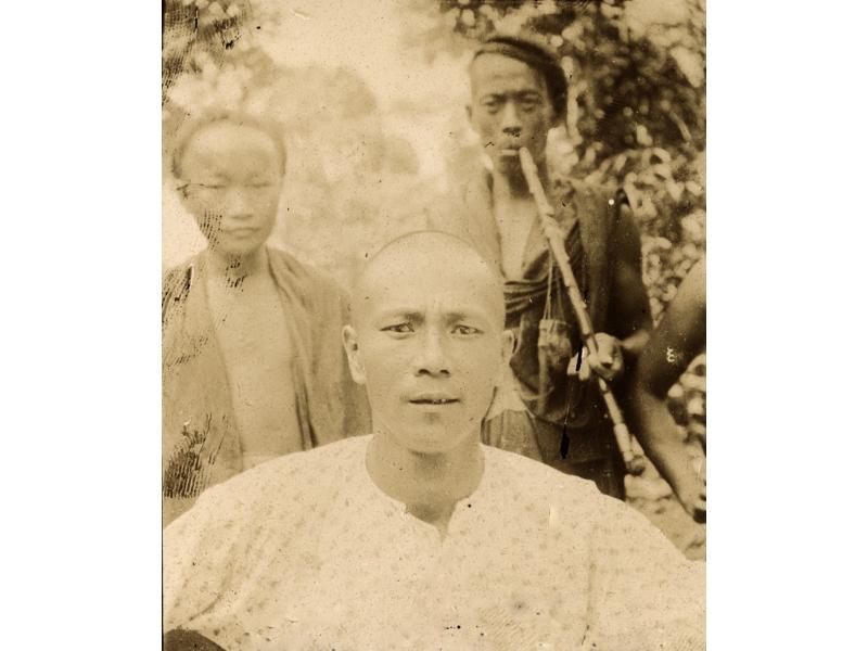 A photo of a man in a white shift. Two men stand behind him, one with his shirt open and the other smoking a long pipe.