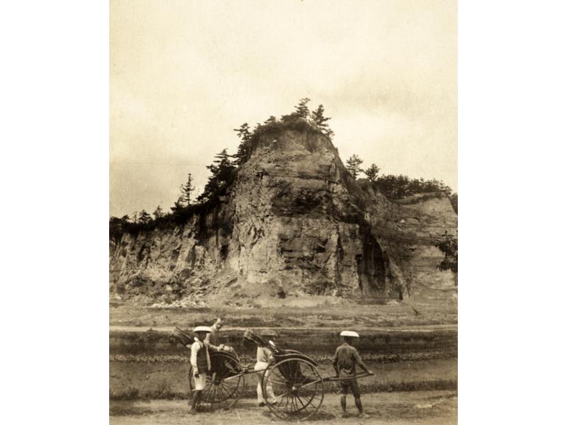 A photo of four people with two rickshaws standing on a road in front of a tall cliff with trees on the top of it.