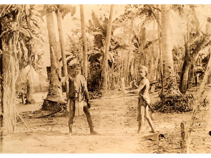 A photo of a man and a boy dressed in the traditional clothing of the Batak tribe. The man is holding a blade and the boy may be as well. They are standing in front of a number of trees.