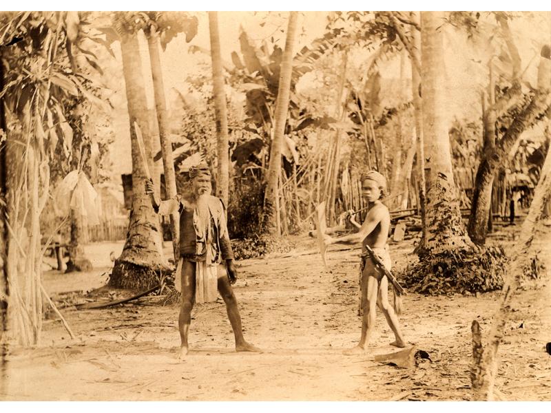 Une photo d’un homme et d’un garçon vêtus des vêtements traditionnels de la tribu Batak. L’homme tient une lame et il se peut que l’enfant en porte également une. Ils sont debout devant quelques arbres.