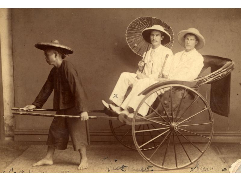 Deux hommes avec des casques coloniaux et des costumes blancs dans un pousse-pousse tiré par un homme de la région. Un foreur tient un parasol et l'autre une canne.