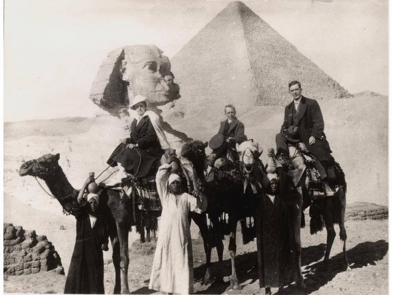 A photo of Mr. and Mrs. Saunders and their son riding camels. There are three local people standing on the ground and holding bottles on their heads. The Sphinx and a pyramid are in the background.